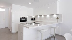 A modern kitchen with white cabinetry, a central island with stools, a built-in oven, and a coffee maker. - Apartment in Playa de Mogán with Sea Views (Mogán)