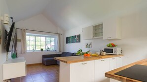 A modern kitchen with a wooden countertop, white cabinets, and a microwave. There is a window with curtains, a gray sofa, and a small plant on the counter. - Bungalow in Maspalomas near Golf & Beach (San Bartolomé de Tirajana)