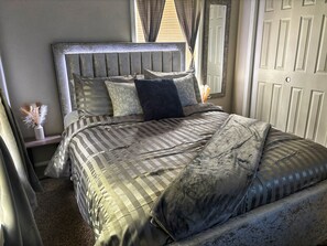 Una cama bien hecha con ropa de cama a rayas y un cabecero, una mesita de noche con un jarrón de flores secas y una puerta con un espejo.