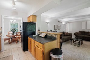 Una cocina moderna con armarios de madera, un refrigerador negro y una pequeña isla. Hay un comedor con una mesa y sillas, y una sala de estar con un sofá y una mesa de centro.