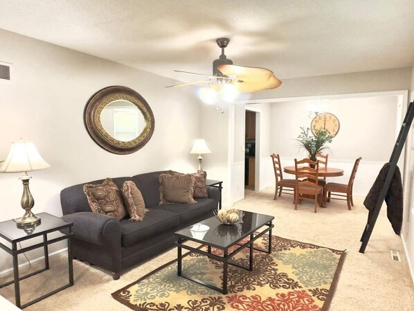 A living room with a sofa, coffee table, lamps, a ceiling fan, a dining table with chairs, and a mirror on the wall.