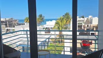 A balcony with a view of the sea, palm trees, and a red car parked on the street.