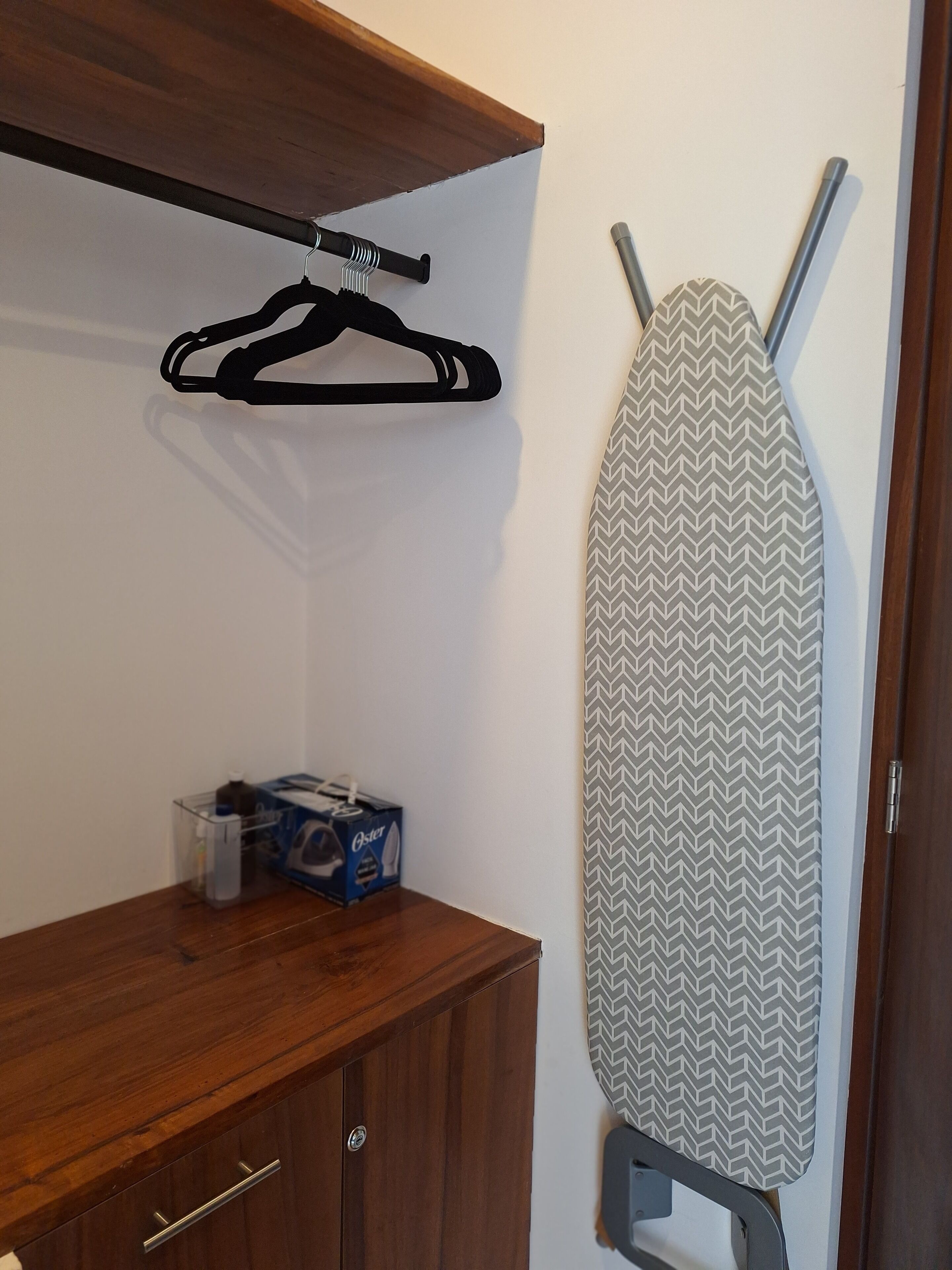A wooden cabinet with a hanging iron on a wall-mounted rack, a box of cleaning supplies, and a patterned ironing board.