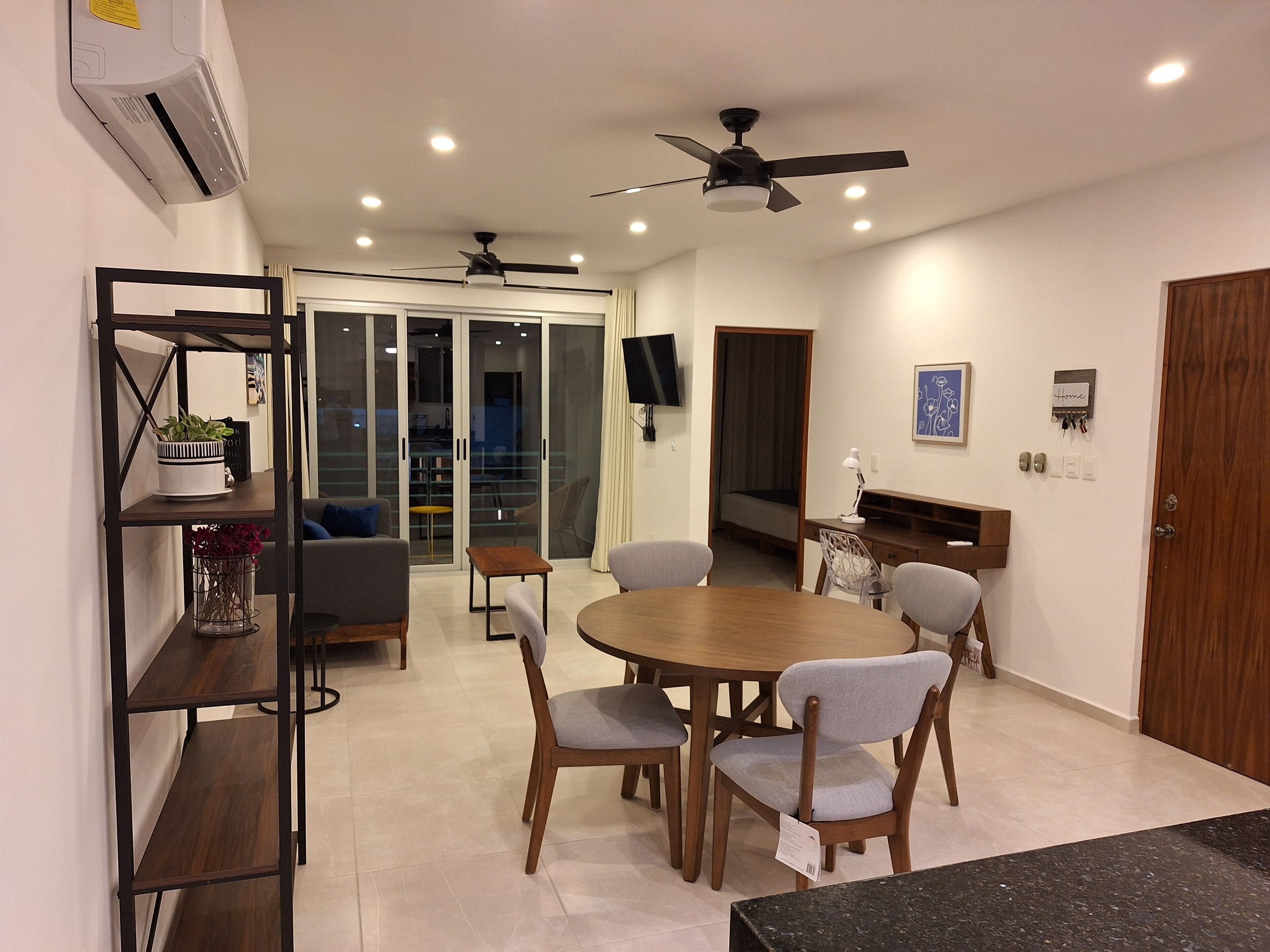 A modern dining area with a round table, six chairs, a wooden bookshelf, and a ceiling fan.