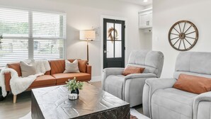 Un salon moderne avec un plafond en bois, un ventilateur de plafond, une grande fenêtre, un canapé en cuir marron, deux fauteuils gris, une table basse en marbre et un tapis en peau de vache.