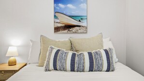 A bedroom with a bed, pillows, a nightstand with a lamp, and a wall-mounted picture of a boat on a beach.