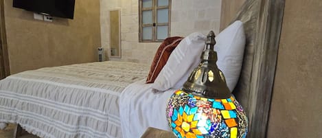 Un dormitorio con cama, televisor montado en la pared y una lámpara decorativa sobre una mesita de noche.