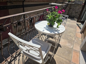 Un balcón con una mesa y una silla blancos, una planta en maceta con flores rosadas y un muro de piedra.