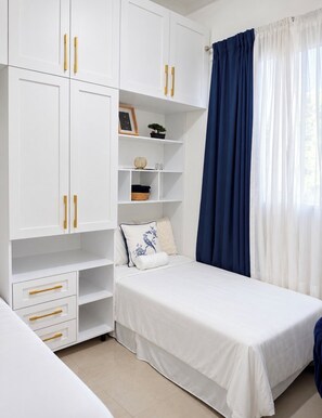 Un dormitorio con cama, armario, estante con adornos y ventana con cortinas.