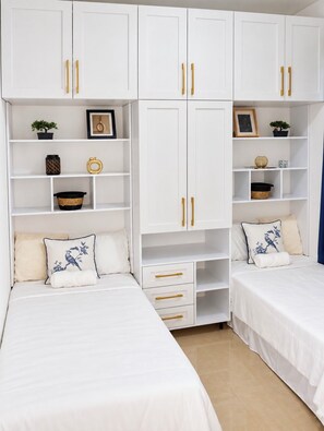 Un dormitorio con dos camas, armarios blancos con tiradores dorados y estanterías con diversos objetos.