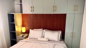 Um quarto bem arrumado com uma cama grande, uma mesa de cabeceira com luminária e guarda-roupas em tons claros com puxadores dourados.