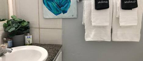Un baño con un lavabo blanco, un cuadro con un diseño abstracto azul y dos toallas con la etiqueta 'Makeup' colgadas en un perchero.