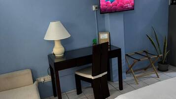 Une pièce comprenant un lit, un bureau avec une lampe, une chaise, un téléviseur fixé au mur et une plante en pot.