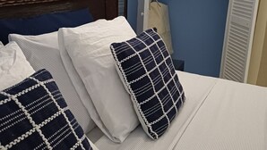 A neatly made bed with a striped bedspread and two decorative pillows. A wooden headboard and a wardrobe with a mirror and folded clothes are visible. - The Palm (Gros Islet)