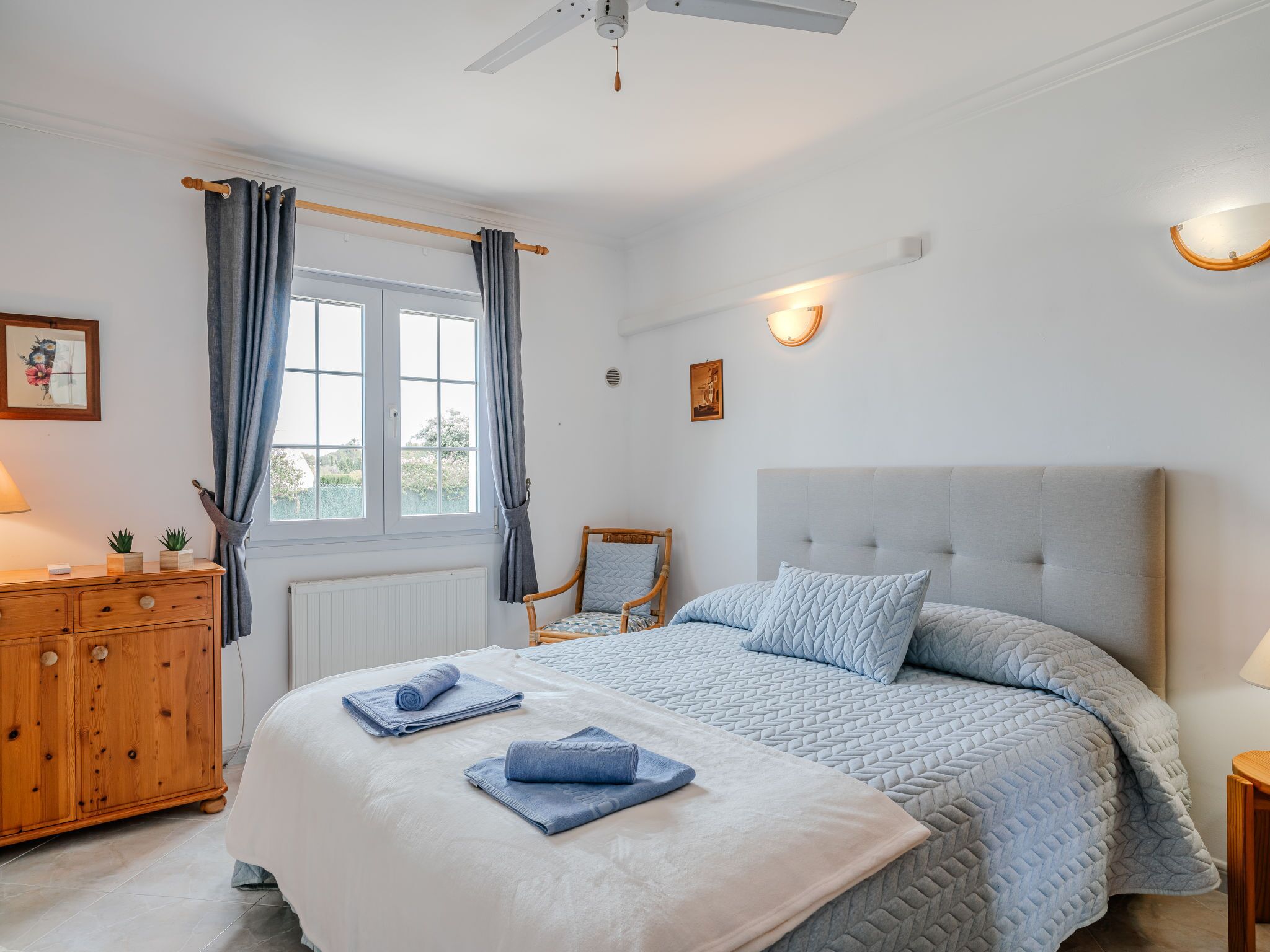 A bedroom with a bed, a wooden dresser, a chair, a window with curtains, and wall-mounted lights.