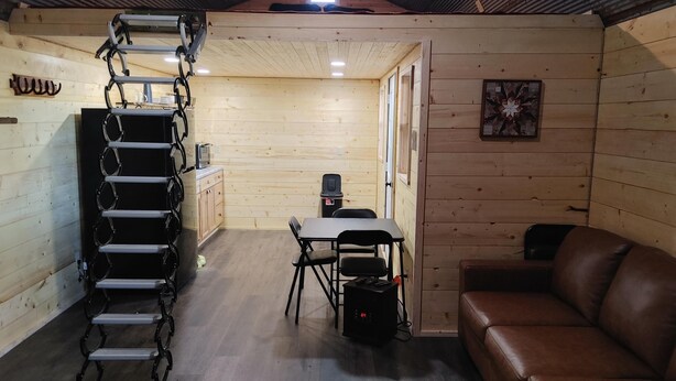 Interior de una cabaña de madera con cama en el altillo, una mesa de comedor con sillas, un sofá de cuero y una pequeña cocina americana.