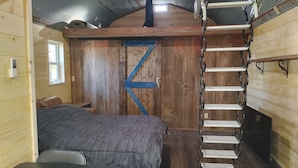 Interior rústico de cabaña con una cama en el altillo, una puerta de madera con una X azul y una escalera de metal.
