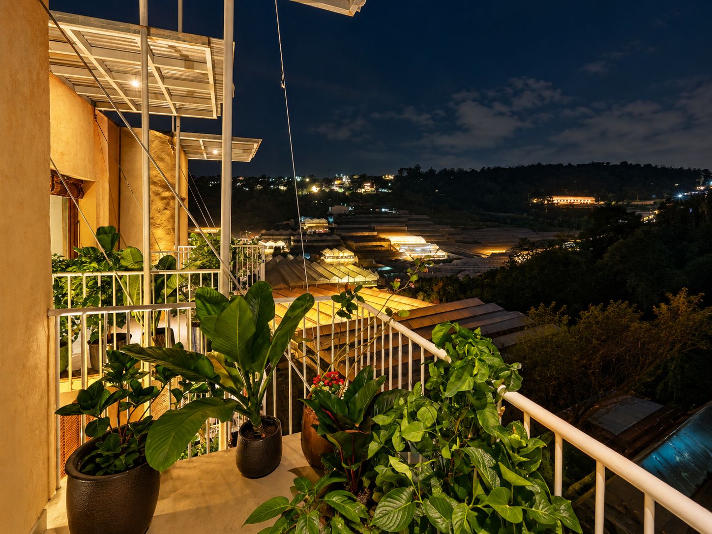 Un balcon orné de plantes en pot, une vue sur un paysage illuminé et un bâtiment en arrière-plan.