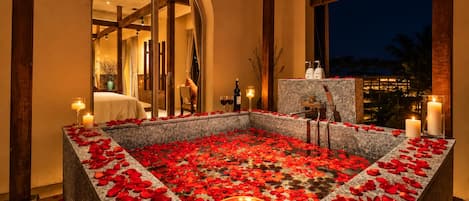 A spa area with a stone bath surrounded by rose petals and candles, a wooden ceiling, and a view of the night sky.