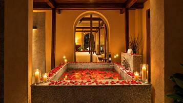 Una piscina con baldosas de piedra, pétalos de rosa y velas, rodeada de pilares y un techo de madera.