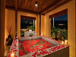 Un área de spa con un jacuzzi de piedra rodeado de pétalos de rosa, techo de madera y vistas a un paisaje urbano distante en la noche.