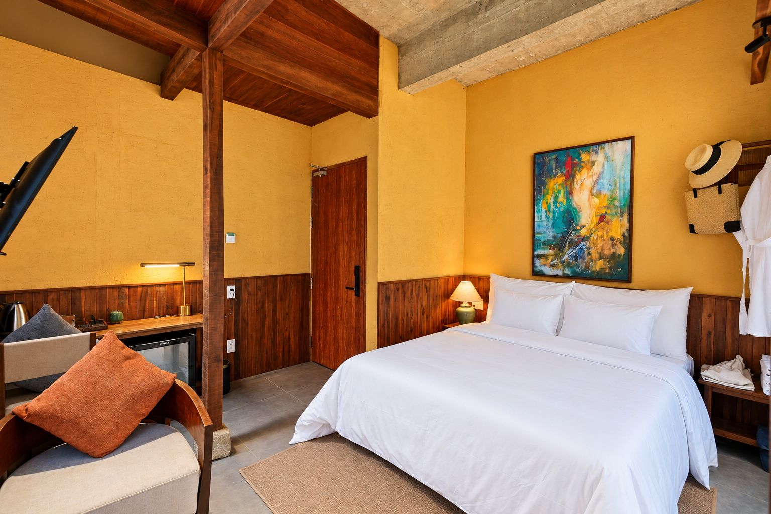 A hotel room with a bed, a desk with a lamp, a painting on the wall, and a hat on the wall.