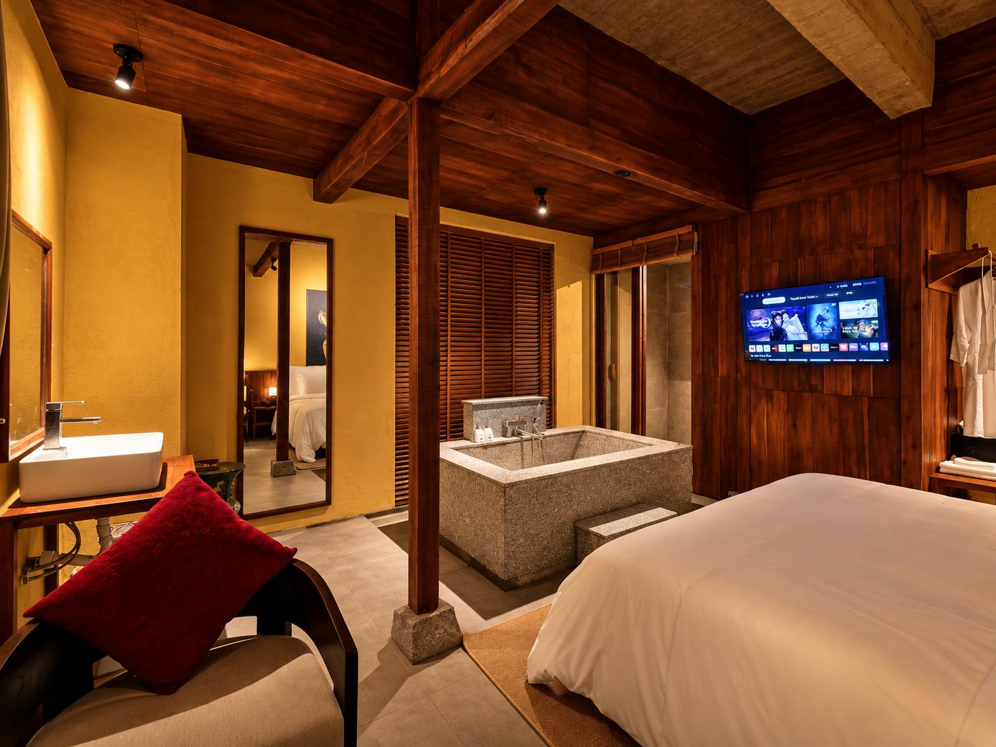 A hotel room with a large bed, a bathroom with a bathtub, a television on the wall, and wooden beams on the ceiling.
