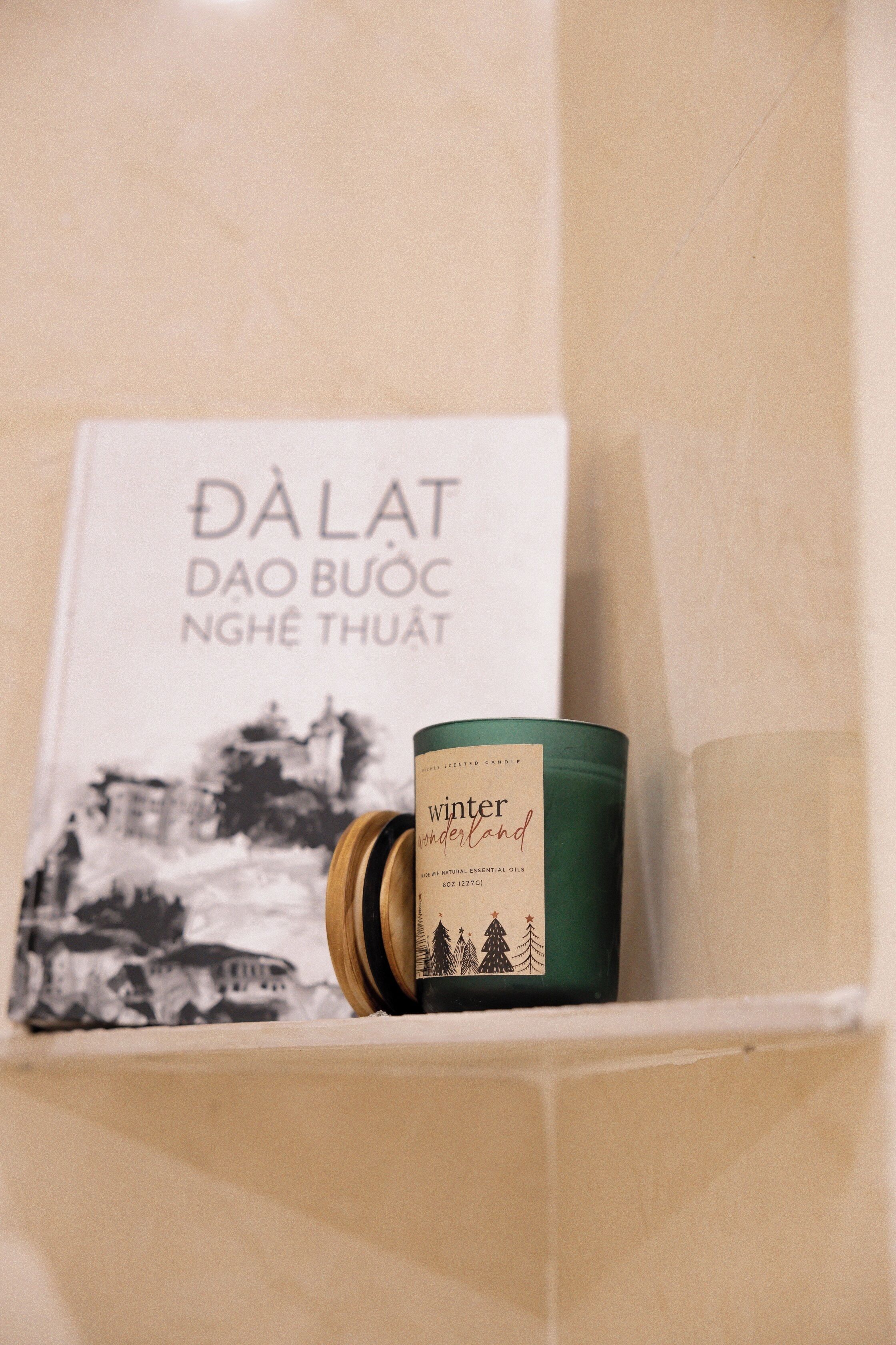 A candle with a label, a book titled "DÂLÂT DAO BUÔC NGHÊ THUẬT," and a wall with a black and white illustration.