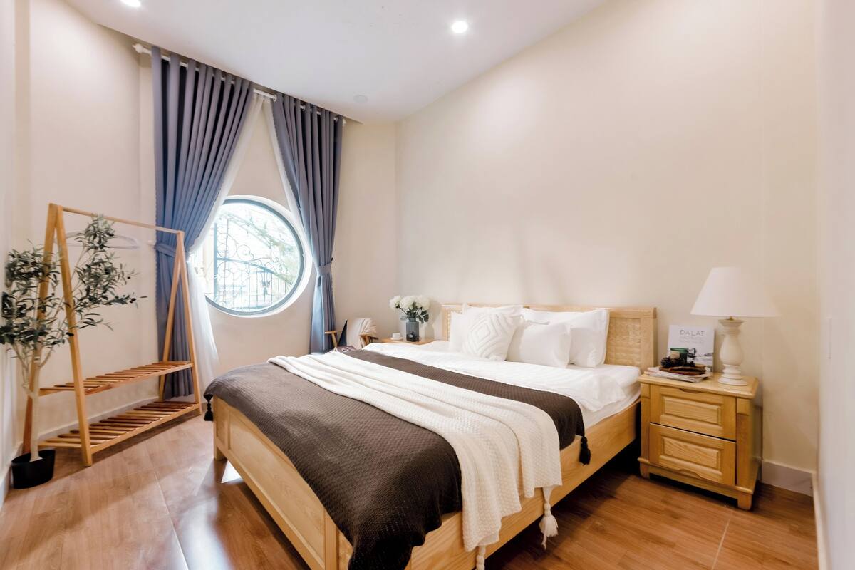 Een slaapkamer met een houten bed, een nachtkastje met een lamp, een raam met gordijnen en een plant op een houten plank.