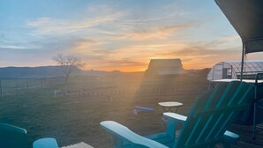 A deck with a wooden table and two turquoise chairs, overlooking a grassy field with a sunset in the background.