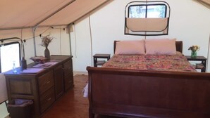 A tented room with a wooden bed, a dresser, a desk with a lamp, and a window.