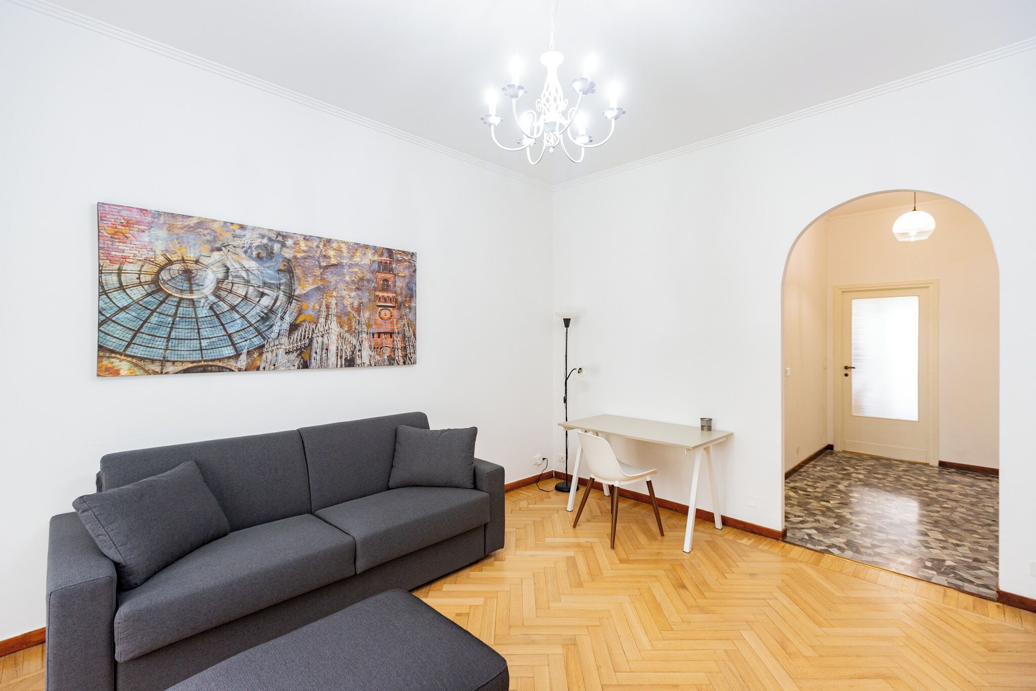 A modern living room with a grey sofa, a dining table with chairs, a chandelier, and a large abstract painting on the wall.