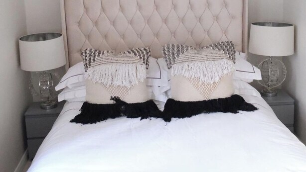 Een net opgemaakt bed met een gestoffeerd hoofdbord, twee bedlampjes en decoratieve kussens.