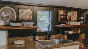 A kitchen with granite countertops, a stainless steel sink, wooden shelves, and various kitchenware.