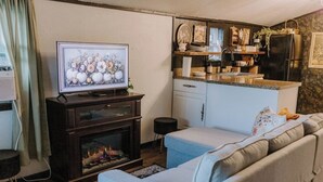 A living room with a fireplace, a television, a sofa, and a kitchen area with a refrigerator and shelves.