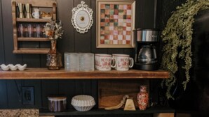 A kitchen with a wooden shelf holding mugs, a coffee maker, and a framed quilt.