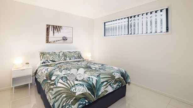 A bedroom with a bed, a nightstand, a wall-mounted picture, and a window with blinds. - Smart Suites Self Check-in Apartments - 18 Darnley (Rocklea)