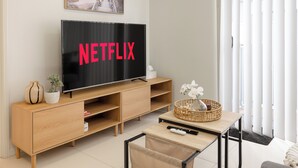 A modern living room with a large flat-screen TV displaying "NETFLIX," a wooden entertainment unit, a small table with a basket, and a chair. - Smart Suites Self Check-in Apartments - 18 Darnley (Rocklea)