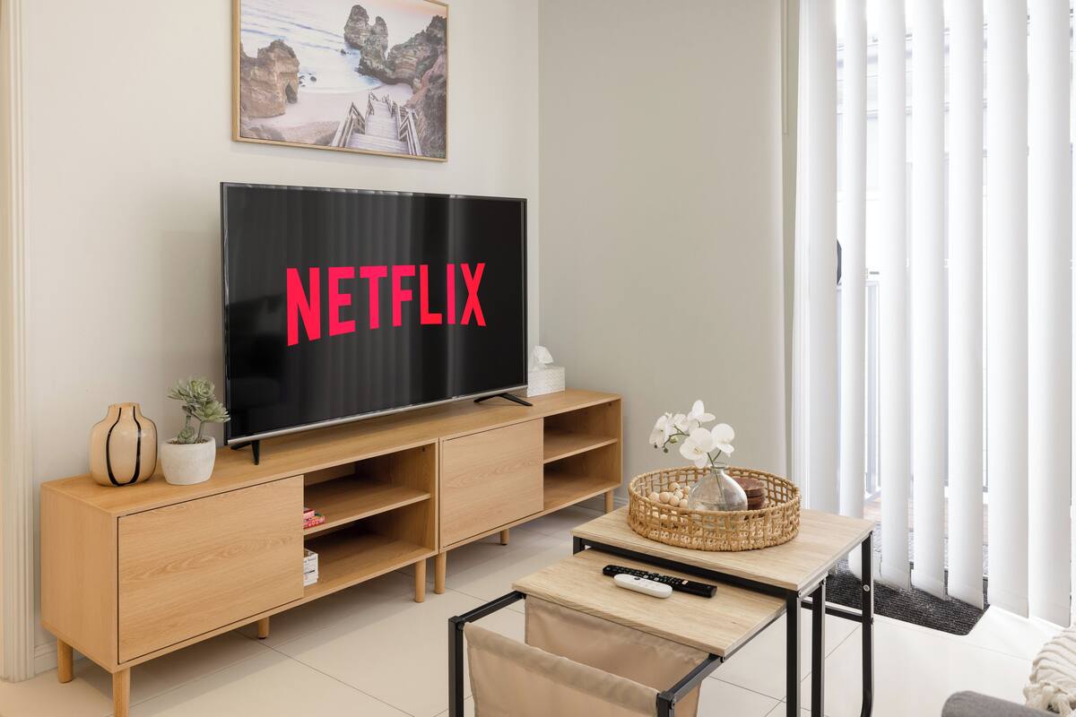 A modern living room with a large flat-screen TV displaying "NETFLIX," a wooden entertainment unit, a small table with a basket, and a chair.