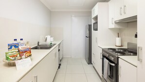 A compact kitchen with white cabinets, a stainless steel refrigerator, a stove, and a microwave. There are various food items and a note on the counter. - Smart Suites Self Check-in Apartments - 18 Darnley (Rocklea)