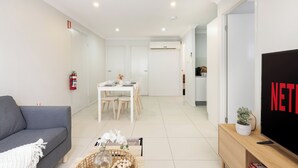 Un salon moderne avec un canapé gris, un coin repas avec une table et des chaises blanches, un téléviseur posé sur un meuble en bois et un extincteur fixé au mur.