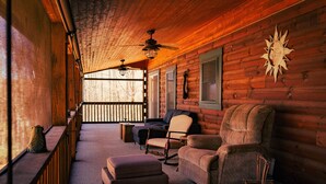 A log cabin interior with a wooden ceiling, two ceiling fans, a large cushioned armchair, a smaller armchair, a bench, and a small table.