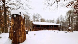 A rustic cabin with a snow-covered roof and a wooden deck, surrounded by trees and a snow-covered ground.
