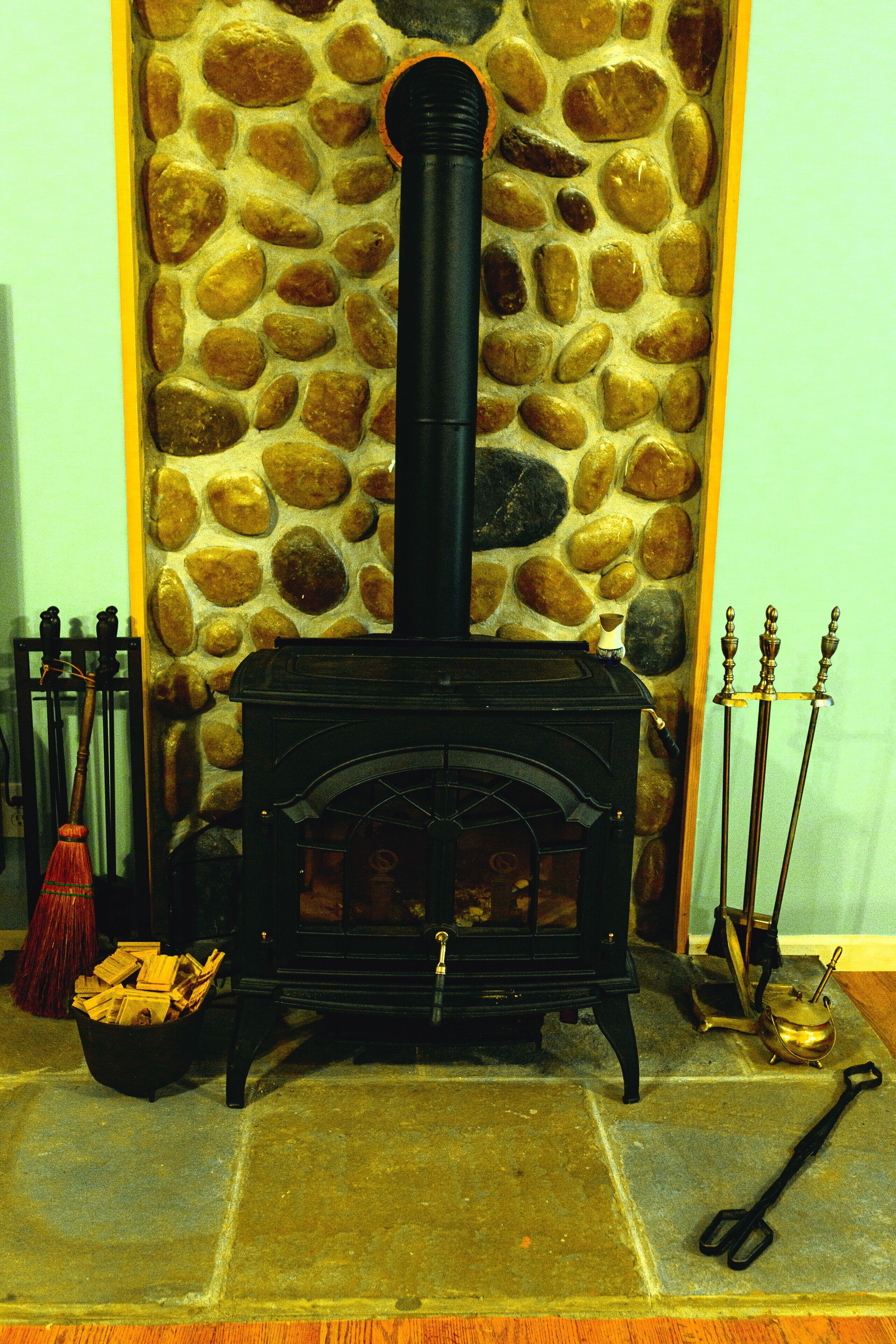 A stone wall with a black wood-burning stove, a broom, a bucket of firewood, and a pair of scissors on the floor.