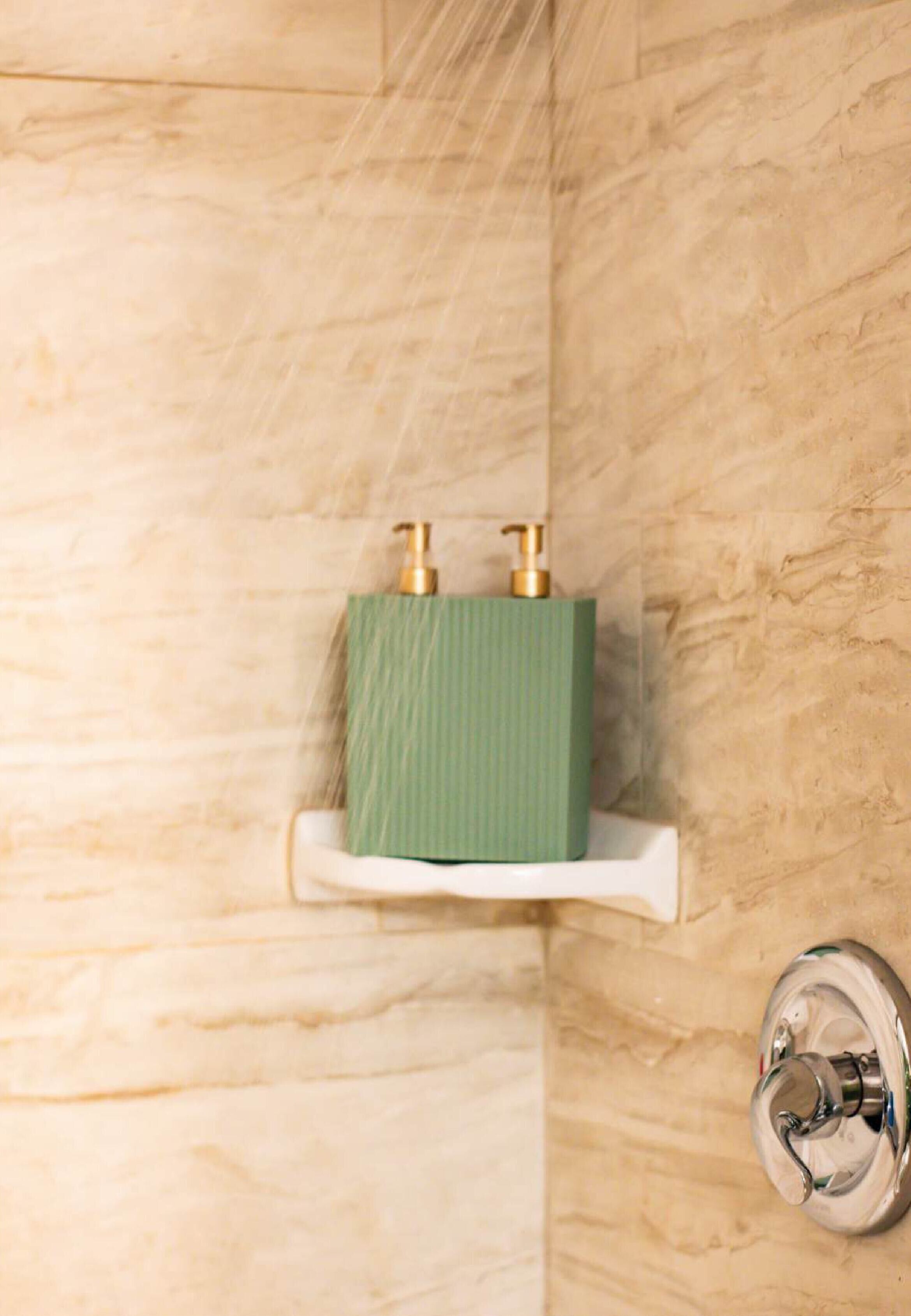 A bathroom with a marble wall, a green soap dispenser on a white shelf, and a chrome shower knob.