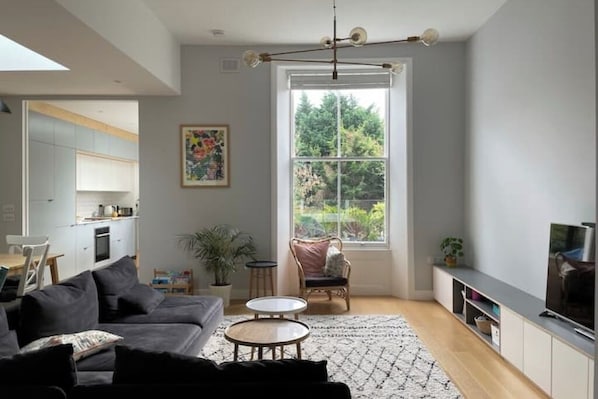 A modern living room with a grey sofa, a coffee table, and a TV on a cabinet. There's a large window with a view of greenery outside.