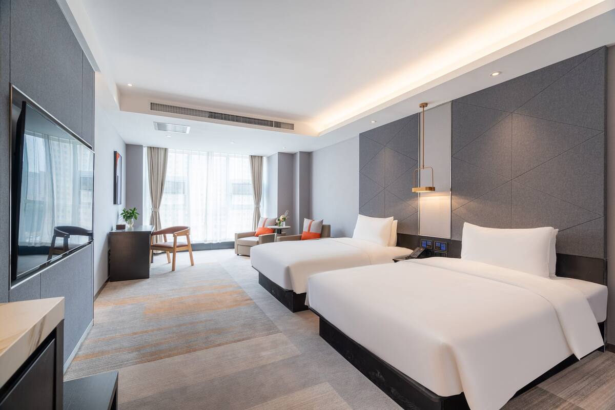 A modern hotel room with two beds, a desk, a chair, a TV, and a large window.