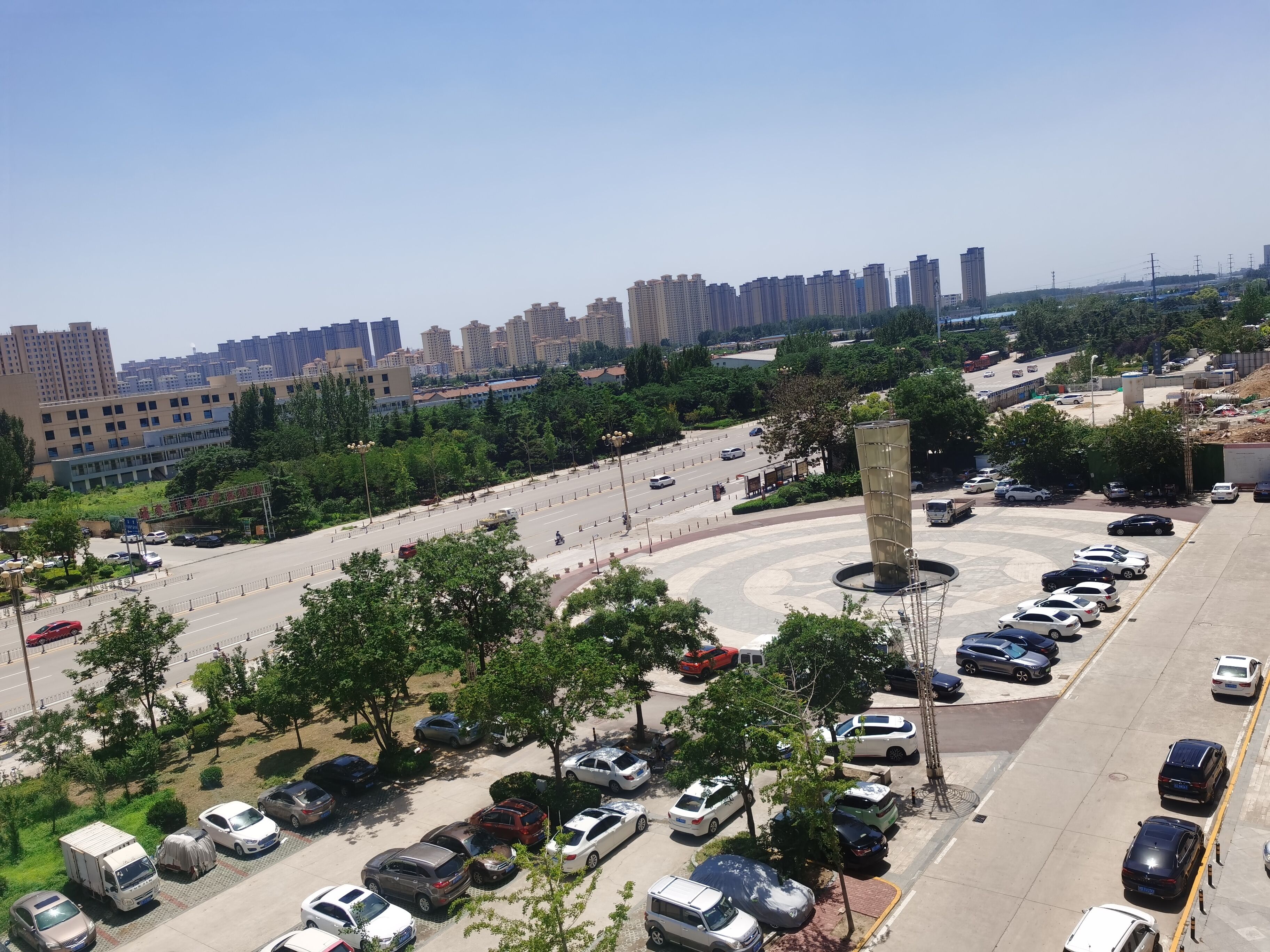 Un estacionamiento con varios autos y algunos camiones, rodeado de árboles y edificios.