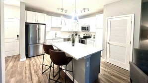 Una cocina moderna con armarios blancos, electrodomésticos de acero inoxidable, una isla central con taburetes y luces colgantes.