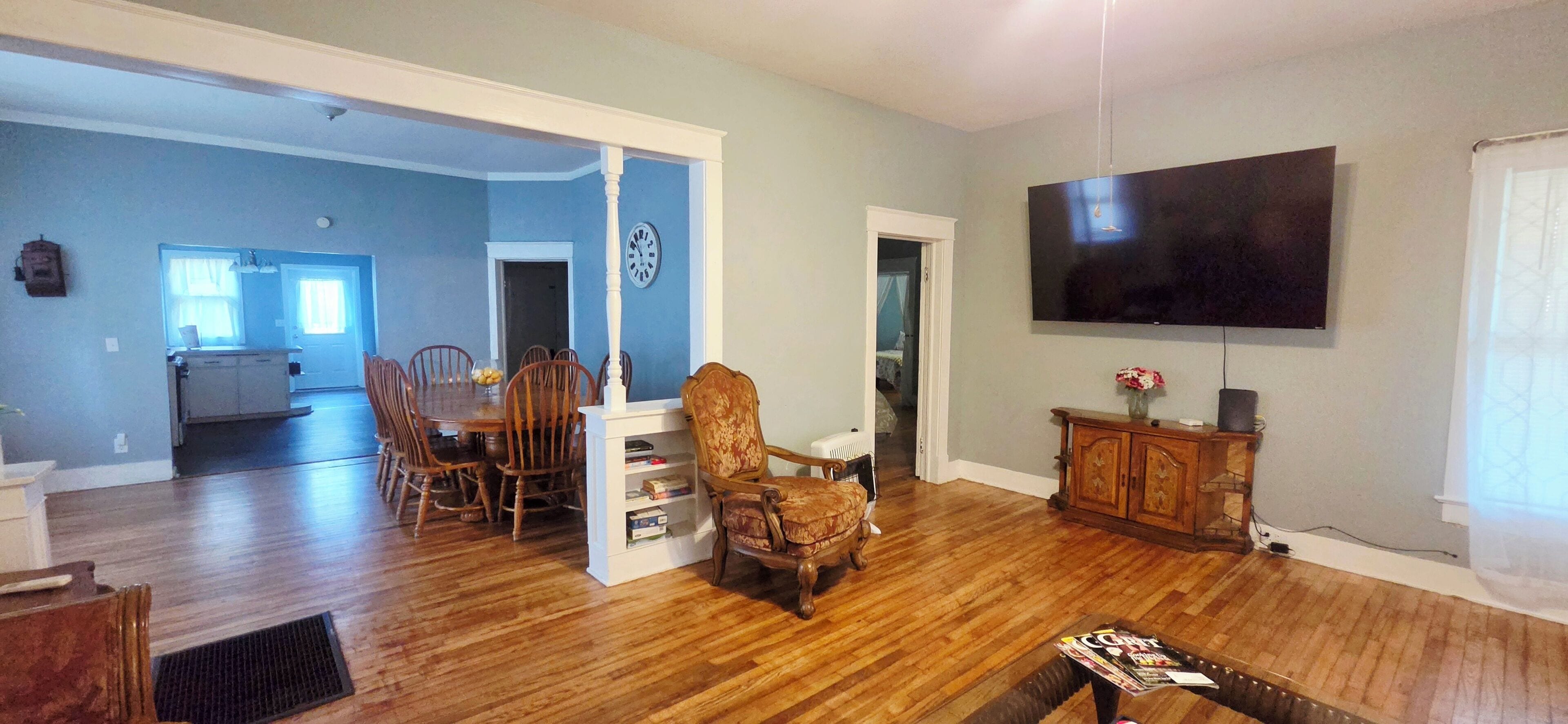 A spacious living room with a dining area, wooden flooring, a flat-screen TV, and a bookshelf.